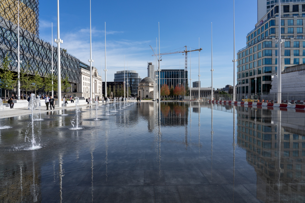 A view of Birmingham's Centenerary Square with fountains in the foreground, library to the left and HSBC UK HQ to the right. Buildings and cranes can be seen in the background.